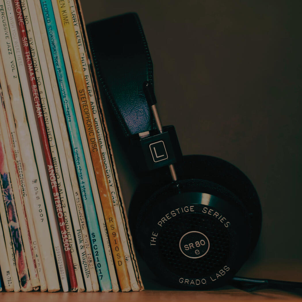 Over-ear headphones resting beside a row of vinyl records on a shelf, with album spines visible in a dimly lit setting.