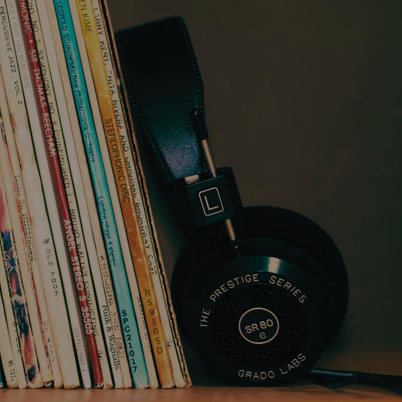 Over-ear headphones resting beside a row of vinyl records on a shelf, with album spines visible in a dimly lit setting.
