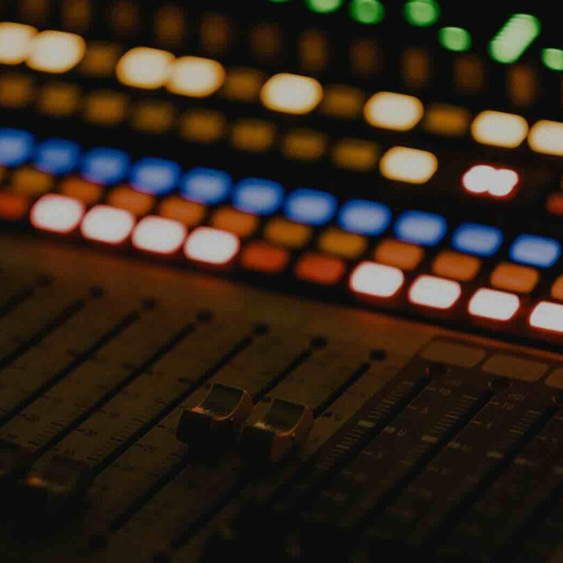 Close-up of an audio mixing console with faders in the foreground and colorful illuminated buttons blurred in the background.