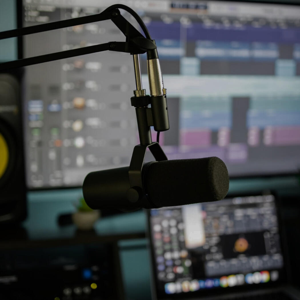 Studio microphone on a boom arm in front of a computer screen showing audio production software, with blurred studio equipment in the background.